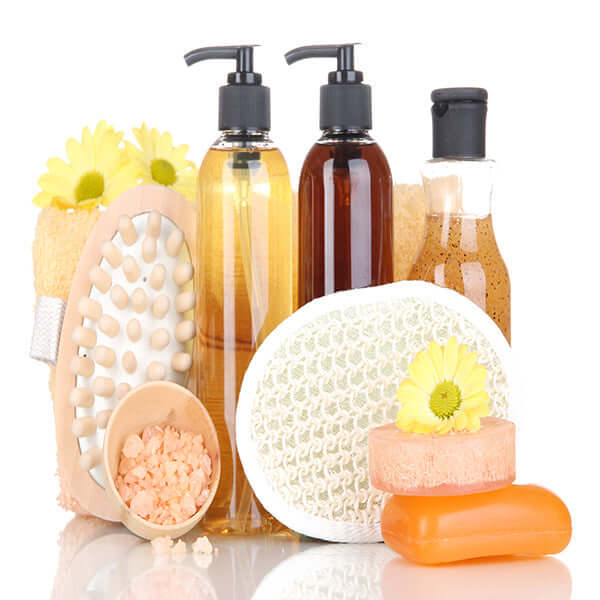Array of beauty products including body wash, scrub brush, loofah, and skincare bottles, adorned with yellow flowers on white background.
